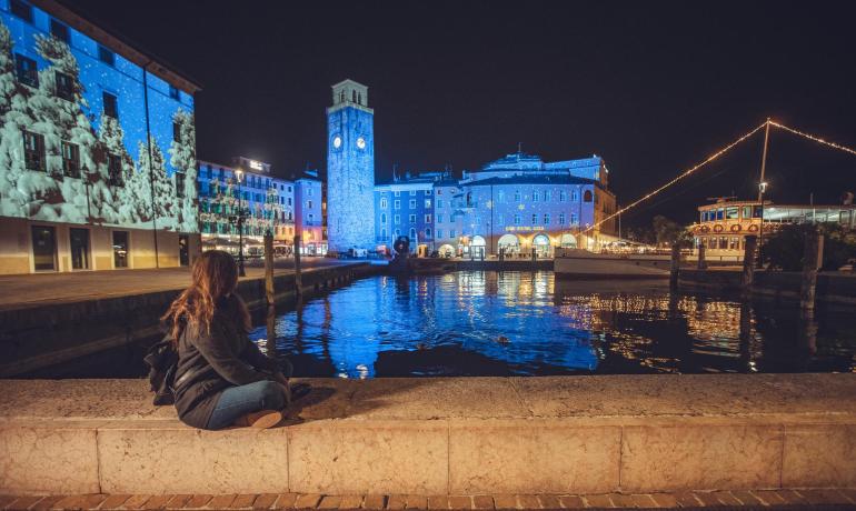 Frau sitzt am See mit beleuchteten Gebäuden bei Nacht.